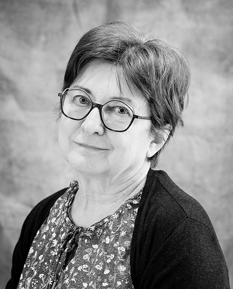 Mustavalkoinen muotokuva iäkkäästä naisesta, jolla on lyhyet hiukset ja pyöreät silmälasit. Hänellä on yllään kukkakuvioinen pusero ja tumma neuletakki. Katse on lempeä ja suora, ja tausta on hillityn utuinen.