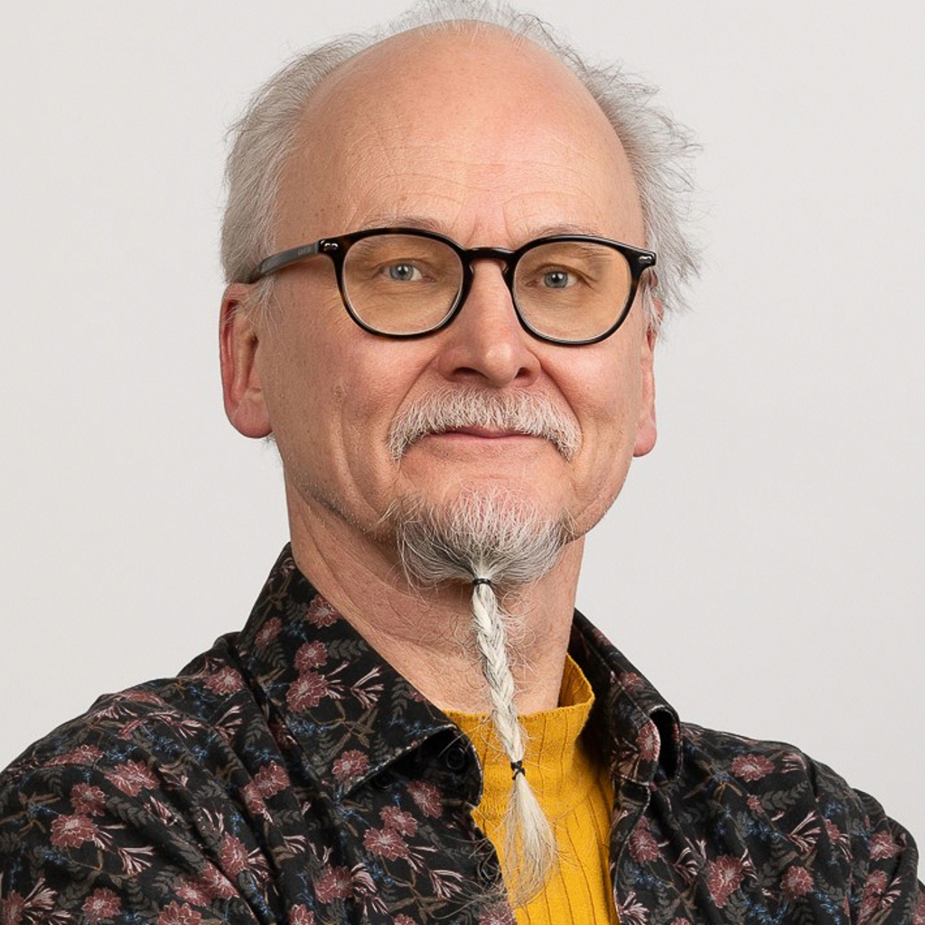 Aikuisen miehen muotokuva. Hänellä on kaljuuntuva pää, lyhyet harmaat hiukset sivuilla ja leuassa pitkähkö vaalea partaletka, joka on letitetty. Hän käyttää pyöreitä mustareunaisia silmälaseja, kukkakuvioista tummasävyistä paitaa ja sen alla keltaista neuletta. Tausta on yksivärinen ja vaalea.