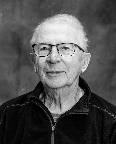 Mustavalkoinen muotokuva iäkkäästä henkilöstä, joka katsoo kameraan lempeästi. Hänellä on silmälasit, lyhyet harmaat hiukset ja yllään vetoketjullinen, tumma pusero. Taustana on pehmeästi sumennettu harmaa studiotausta.