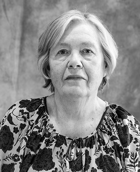 Mustavalkoinen muotokuva iäkkäästä naisesta, jolla on hartioille ulottuvat, hieman lainehtivat hiukset. Hänellä on kukkakuvioinen pusero ja neutraali ilme. Tausta on pehmeästi epätarkka, harmaasävyinen.