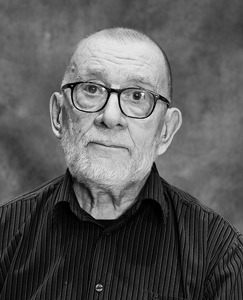 Mustavalkoinen muotokuva iäkkäästä henkilöstä, jolla on lyhyet hiukset ja parta. Hänellä on pyöreät silmälasit ja raidallinen kauluspaita. Henkilö katsoo kameraan rauhallisesti. Taustana on harmaa studiotausta.