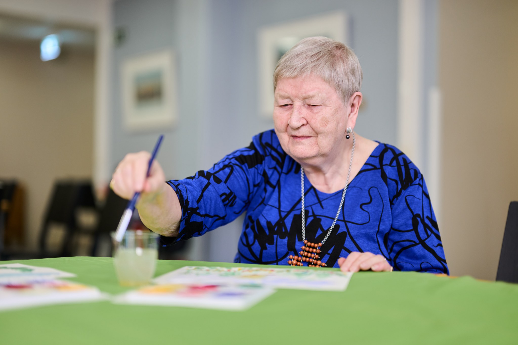 Iäkäs nainen maalaa paperille siveltimellä pöydän ääressä. Hän keskittyy työhönsä ja käyttää sinistä paitaa sekä kaulakorua. Pöydällä on vihreä pöytäliina ja maalausvälineitä. Taustalla näkyy muita ihmisiä ja pöytiä.
