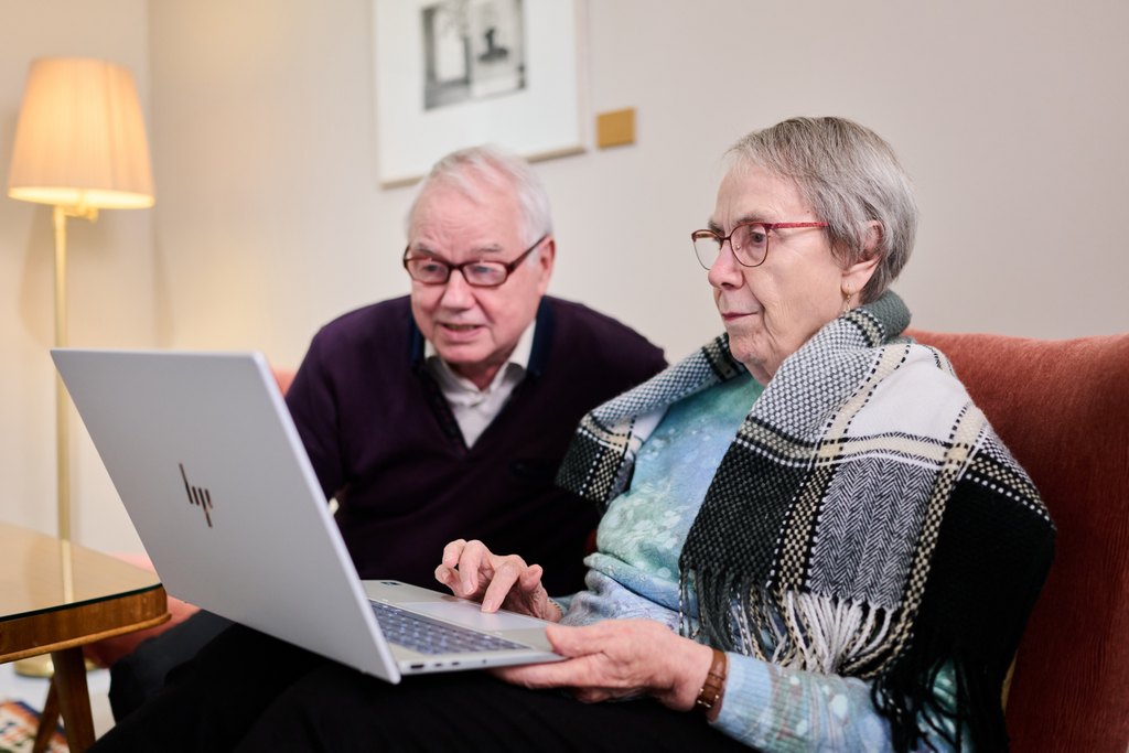 Kaksi iäkästä henkilöä istuu vierekkäin sohvalla ja katsoo kannettavan tietokoneen ruutua. Oikealla istuva henkilö käyttää konetta ja vasemmalla istuva seuraa kiinnostuneena vierestä. Molemmilla on silmälasit, ja taustalla näkyy lamppu ja seinätaidetta. Tunnelma on rauhallinen ja keskittynyt.