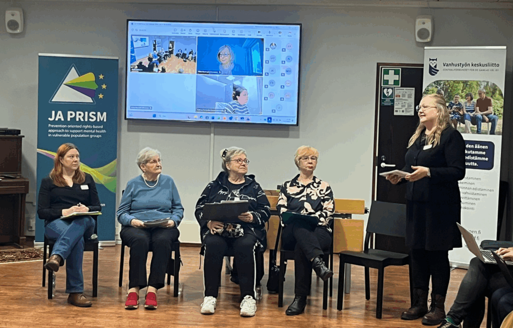 Paneelikeskustelu sisätilassa. Viisi henkilöä istuu tuoleilla rivissä ja yksi henkilö seisoo edessä puhumassa. Taustalla näkyy näyttö, jossa osa osallistujista on mukana etäyhteydellä. Tilassa on roll-upit, joissa näkyvät JA PRISM -hankkeen ja Vanhustyön keskusliiton logot.