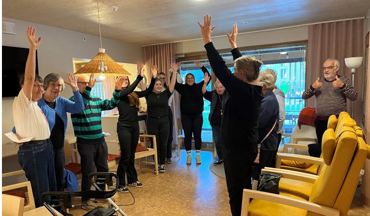 Ryhmä eri-ikäisiä ihmisiä seisoo ringissä valoisassa oleskelutilassa ja tekee yhteisiä venyttely- tai jumppaliikkeitä kädet ylös kohotettuina. Mukana on sekä iäkkäitä että nuorempia osallistujia, ja tunnelma on iloinen ja kannustava. Taustalla näkyy ikkunoita, verhoja ja kodikkaita kalusteita, kuten keltaisia nojatuoleja ja rottinkinen kattovalaisin.