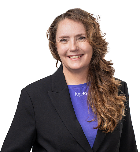 Hymyilevä nainen seisoo valkoista taustaa vasten. Hänellä on pitkät ruskeat, kevyesti laineilevat hiukset ja yllään tummansininen paita sekä musta bleiseri. Nainen katsoo suoraan kameraan ystävällinen ilme kasvoillaan.