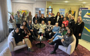 Ryhmäkuva sisätilassa. Eri-ikäisiä ihmisiä istuu ja seisoo kahden sohvan ympärillä rennossa asetelmassa. Etualalla on pieni pöytä ja taustalla näkyy JA PRISM -hankkeen roll-up. Tunnelma on lämmin ja yhteisöllinen.