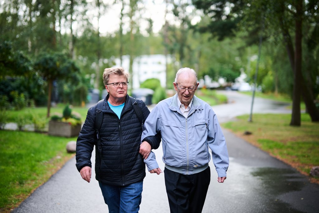 Kaksi iäkästä ihmistä kävelee käsikynkässä puistomaisella kävelytiellä. Ympärillä on puita ja vihreää, ja tunnelma on rauhallinen ja lämmin