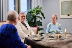 Kolme iäkästä henkilöä istuu pöydän ääressä yhteisessä tilassa ja keskustelee. Pöydällä on kahvikuppeja, tarjoiluastioita ja kynttilä. Taustalla näkyy viherkasvi ja seinällä taulu. Tunnelma on rauhallinen, lämmin ja yhdessäoloon keskittynyt.