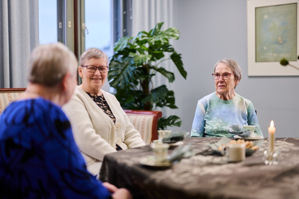 Kolme iäkästä henkilöä istuu pöydän ääressä yhteisessä tilassa ja keskustelee. Pöydällä on kahvikuppeja, tarjoiluastioita ja kynttilä. Taustalla näkyy viherkasvi ja seinällä taulu. Tunnelma on rauhallinen, lämmin ja yhdessäoloon keskittynyt.