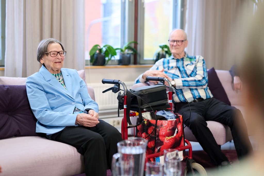 Kaksi iäkästä henkilöä istuu vierekkäin vaalealla sohvalla yhteisessä tilassa. Vasemmalla istuva henkilö hymyilee rauhallisesti ja katsoo eteenpäin, oikealla istuva henkilö nojaa taaksepäin ja katsoo toista kohti. Heidän välissään on punainen rollaattori, jonka edessä on kuvioitu laukku. Taustalla näkyy ikkuna, verhot ja viherkasveja. Tunnelma on levollinen ja kodinomainen.