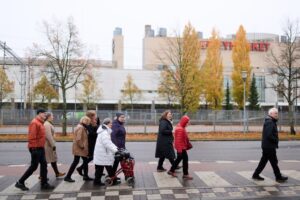 Ryhmä eri-ikäisiä aikuisia ja iäkkäitä ihmisiä ylittää suojatietä syksyisessä kaupunkiympäristössä. Yhdellä osallistujalla on rollaattori, ja taustalla näkyy puita ja rakennuksia.