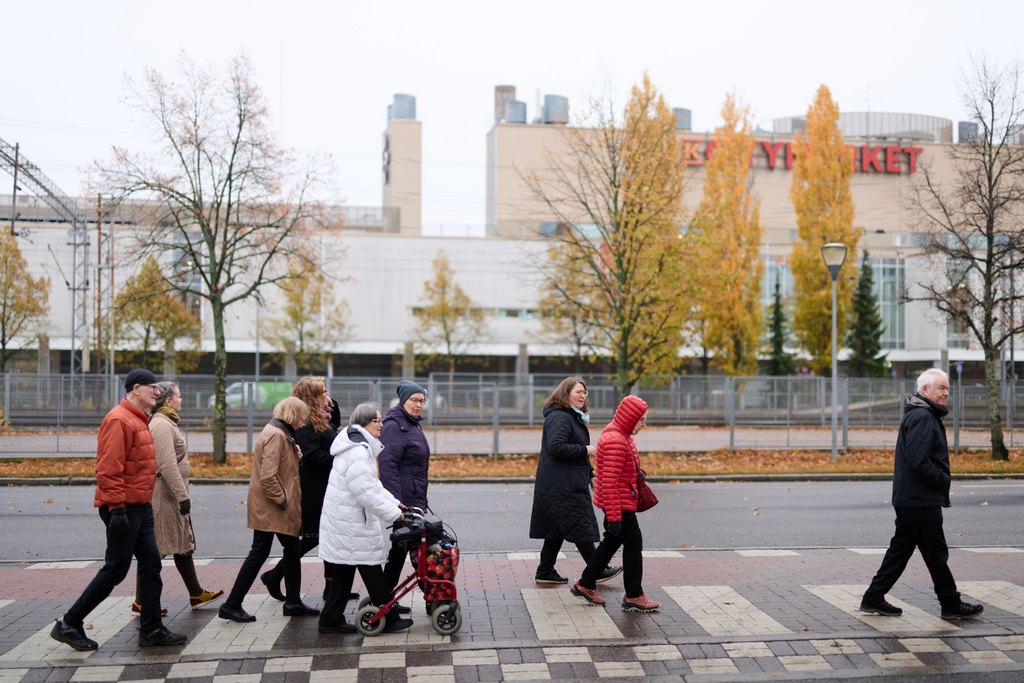 Ryhmä eri-ikäisiä aikuisia ja iäkkäitä ihmisiä ylittää suojatietä syksyisessä kaupunkiympäristössä. Yhdellä osallistujalla on rollaattori, ja taustalla näkyy puita ja rakennuksia.