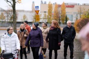 Ryhmä iäkkäitä ihmisiä kävelee yhdessä jalkakäytävällä kaupunkiympäristössä syksyisenä päivänä. Yhdellä henkilöllä on rollaattori, muilla on ulkovaatteet ja käsilaukkuja. Taustalla näkyy katu, liikennemerkkejä ja rakennuksia. Tunnelma on rauhallinen ja yhteisöllinen.