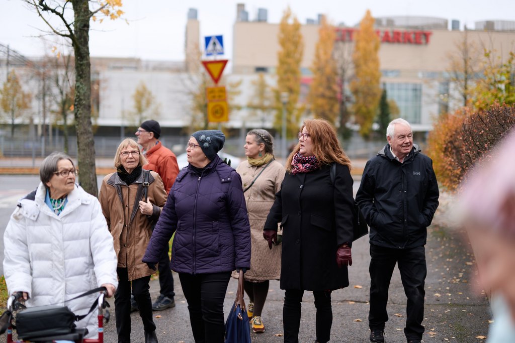Ryhmä iäkkäitä ihmisiä kävelee yhdessä jalkakäytävällä kaupunkiympäristössä syksyisenä päivänä. Yhdellä henkilöllä on rollaattori, muilla on ulkovaatteet ja käsilaukkuja. Taustalla näkyy katu, liikennemerkkejä ja rakennuksia. Tunnelma on rauhallinen ja yhteisöllinen.