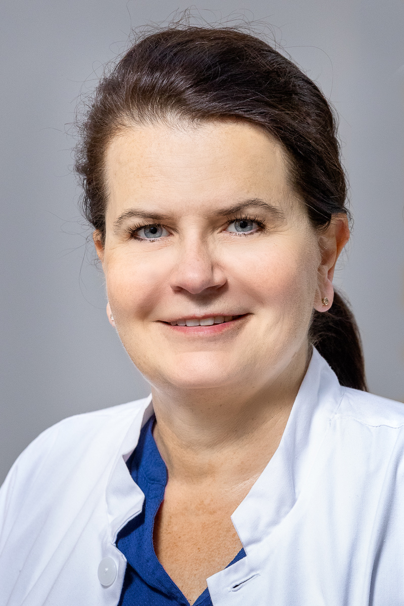 Naisen kasvokuva neutraalia taustaa vasten. Hänellä on tumma hius, valkoinen työtakki ja sininen paita. Ilme on rauhallinen ja ystävällinen.