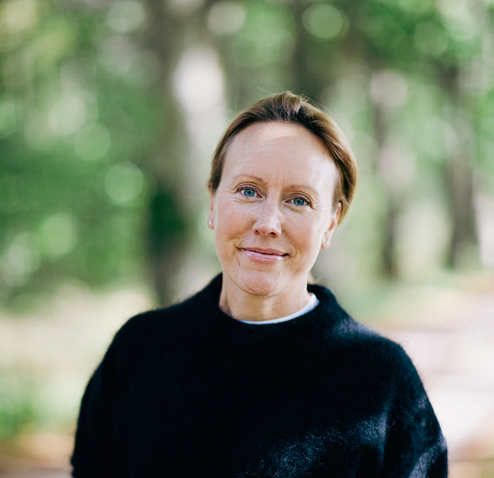 En person står utomhus i en skogsmiljö och ser in i kameran. Bakgrunden är grön och mjukt suddig, med en lugn och naturinspirerad stämning.