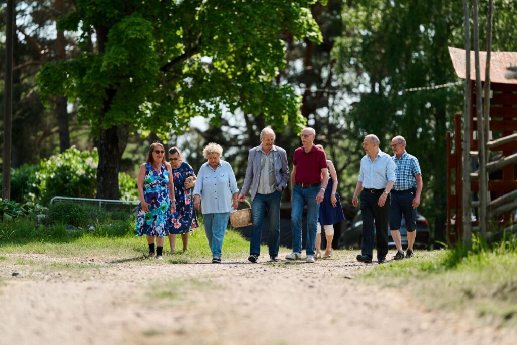 En grupp äldre personer går tillsammans i ett somrigt landskap