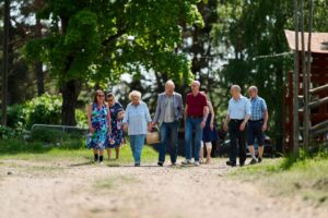 Ryhmä ikääntyneitä ihmisiä kävelee yhdessä aurinkoisena kesäpäivänä hiekkatietä pitkin vehreässä maaseutuympäristössä. He keskustelevat ja viettävät aikaa yhdessä.