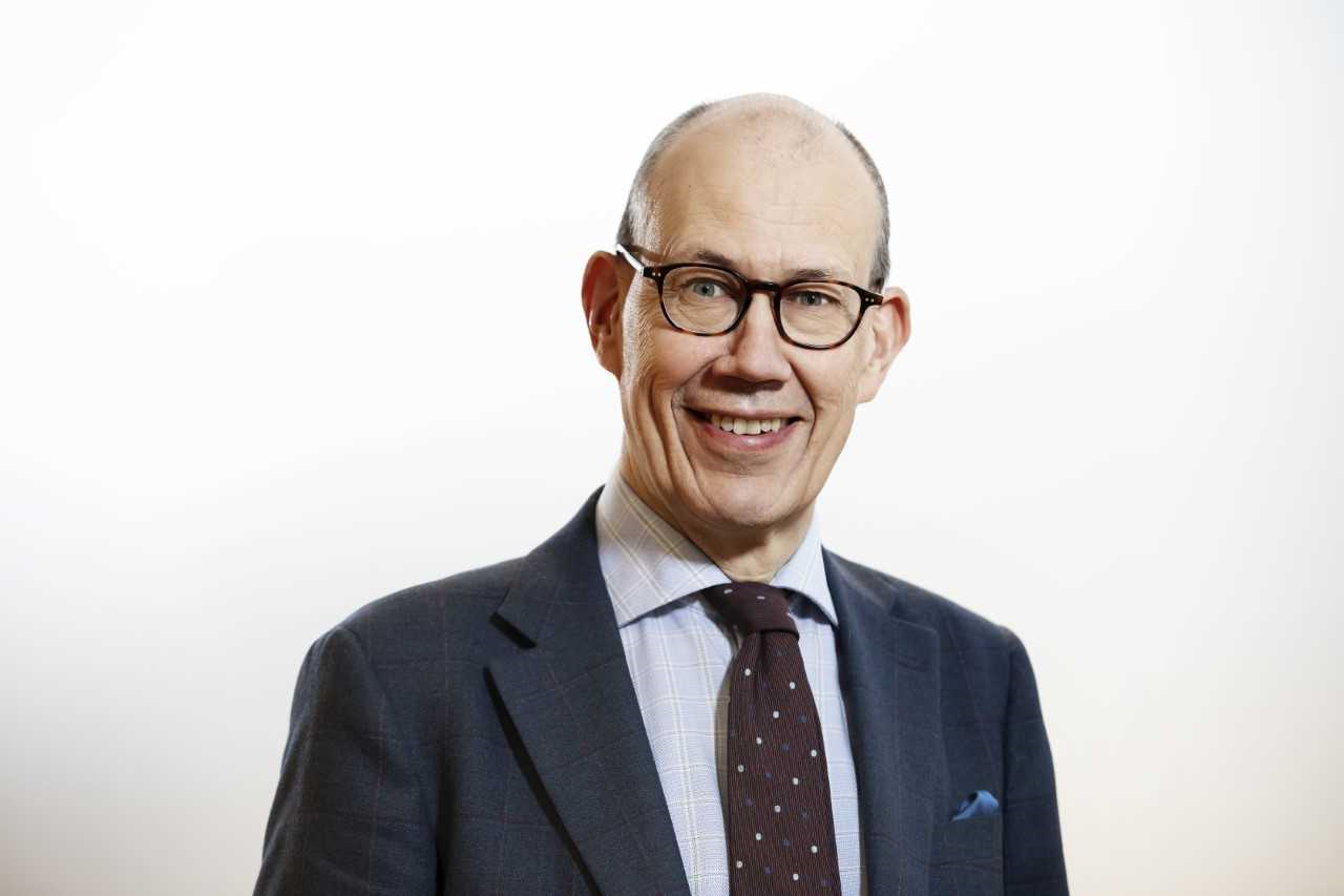 Miehen muotokuva vaaleaa taustaa vasten. Hänellä on silmälasit, puku, solmio ja taskuliina. Hän hymyilee suoraan kameraan.