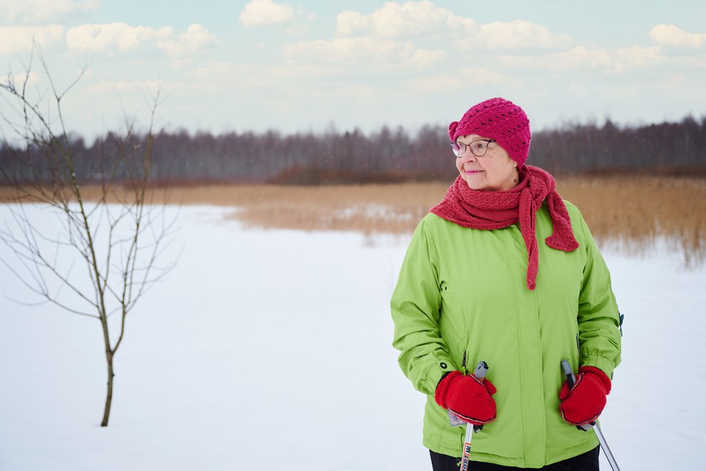 Iäkäs henkilö seisoo talvisessa maisemassa lumisella pellolla. Hänellä on vihreä ulkoilutakki, punainen pipo ja huivi sekä kävelysauvat käsissään. Taustalla näkyy puita ja avaraa talvista luontoa.