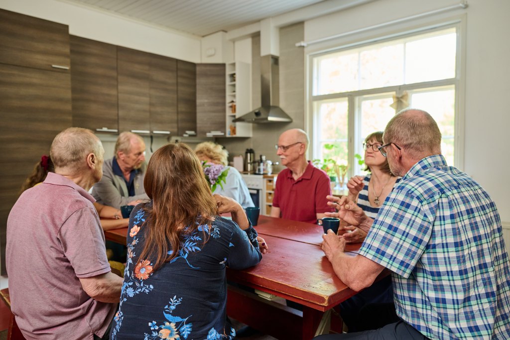 En grupp äldre personer sitter runt ett köksbord och samtalar över kaffekoppar. I bakgrunden syns ett hemtrevligt kök och ett fönster med dagsljus.