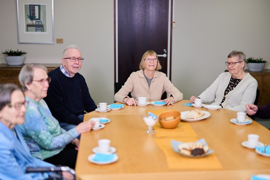 En grupp äldre personer sitter runt ett bord med kaffekoppar och servering. De samtalar tillsammans inomhus i en lugn och gemenskaplig miljö.
