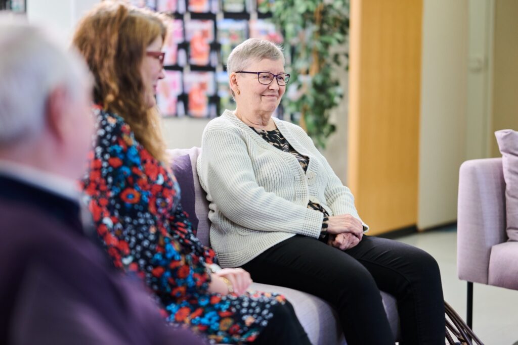 Iäkäs nainen istuu tuolilla ryhmätilanteessa ja hymyilee rauhallisesti. Hänen vieressään istuu toinen henkilö, ja taustalla näkyy esiteteline sekä viherkasvi. Tunnelma on lämmin ja osallistava.