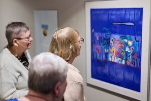 Kolme iäkästä henkilöä tarkastelee seinälle ripustettua värikästä taideteosta sisätilassa. He seisovat vierekkäin ja katsovat teosta keskittyneesti.