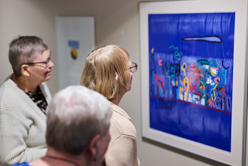 Kolme iäkästä henkilöä tarkastelee seinälle ripustettua värikästä taideteosta sisätilassa. He seisovat vierekkäin ja katsovat teosta keskittyneesti.