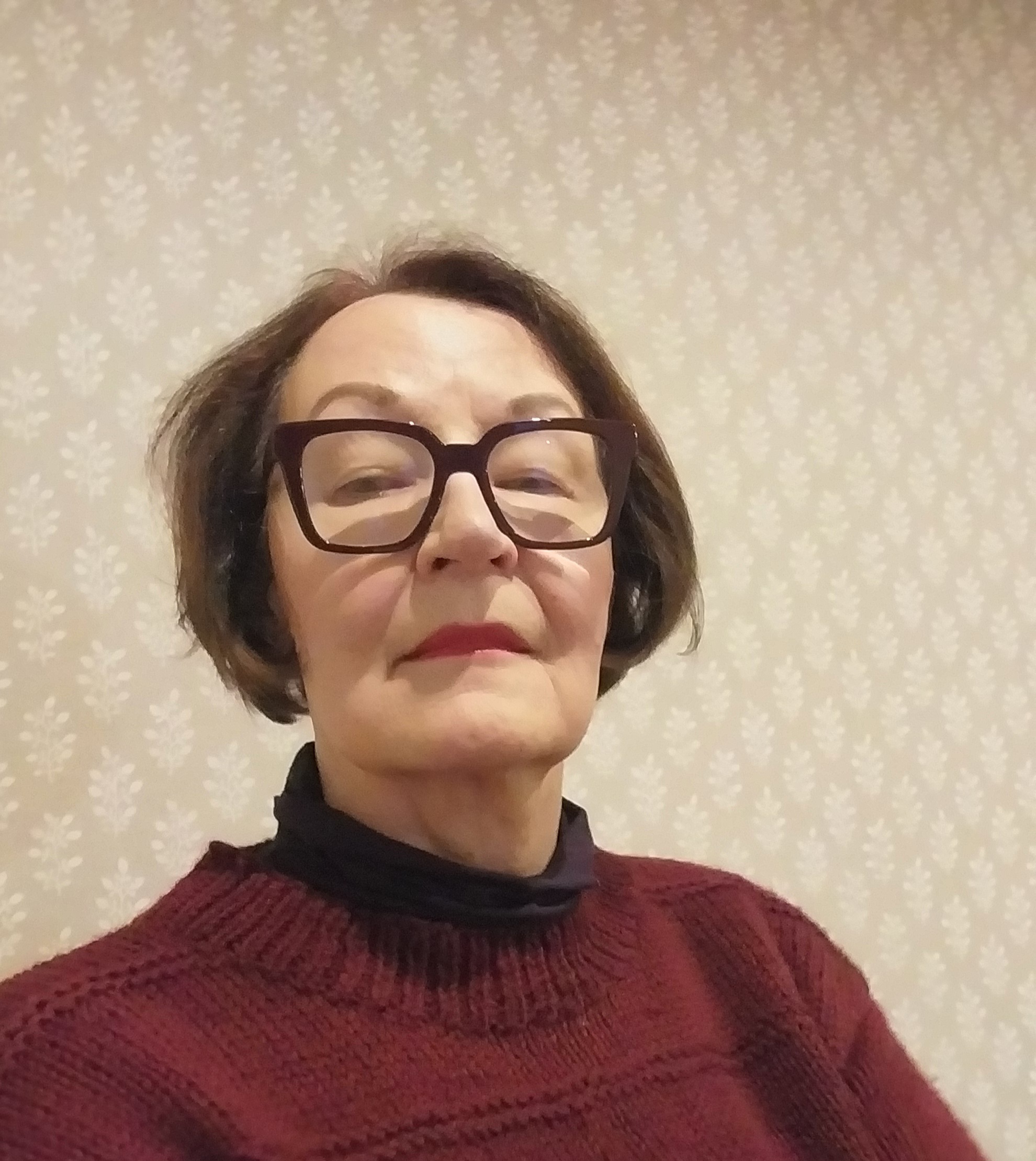 Henkilökuva sisätiloissa: lyhyttukkainen henkilö katsoo kameraan. Hänellä on tummat silmälasit, punainen neule ja tumma poolopaita. Taustalla vaalea, hillitysti kuvioitu seinä.