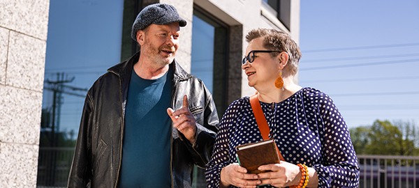 Kaksi aikuista henkilöä kävelee ulkona rakennuksen edustalla aurinkoisena päivänä. He keskustelevat iloisesti keskenään. Toinen osoittaa sormellaan puhuessaan, ja toisella on kädessään muistikirja. Tunnelma on lämmin ja vuorovaikutteinen.