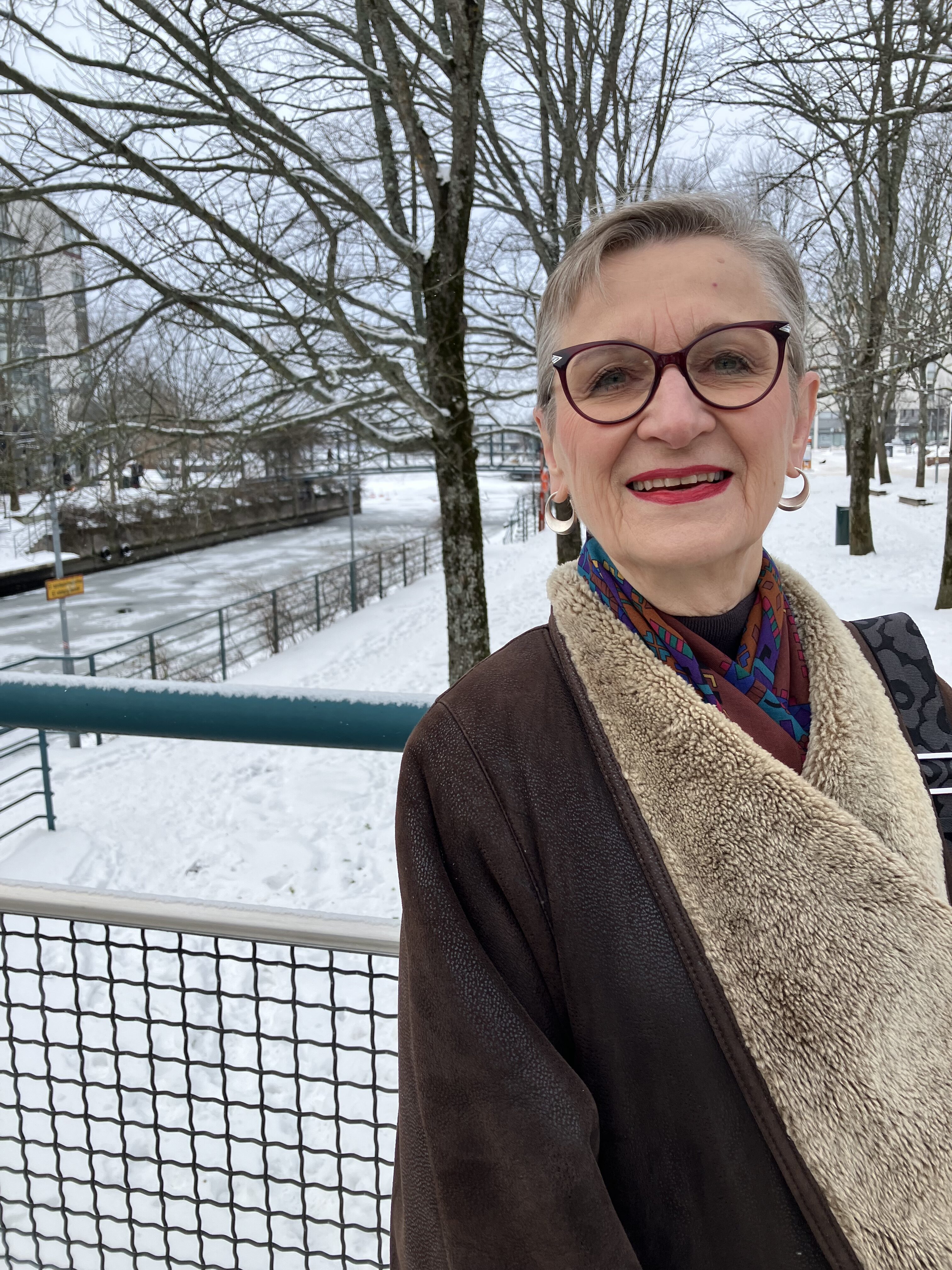 Henkilökuva ulkona talvisessa ympäristössä: hymyilevä lyhyttukkainen henkilö seisoo sillalla. Hänellä on silmälasit, ruskea takki ja värikäs huivi. Taustalla näkyy luminen maisema, puita ja kanava.