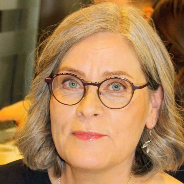 Lyhythiuksinen, harmaahiuksinen nainen katsoo suoraan kameraan. Hänellä on pyöreät silmälasit ja hillitty meikki. Tausta on lämminsävyinen ja hieman epätarkka.