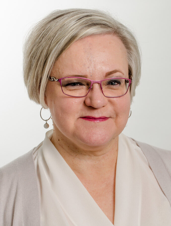 Vaaleahiuksinen nainen katsoo kameraan ja hymyilee hienovaraisesti. Hänellä on lyhyt, vaalea kampaus, pinkkikehyksiset silmälasit ja vaalea pusero. Tausta on neutraali ja vaalea.