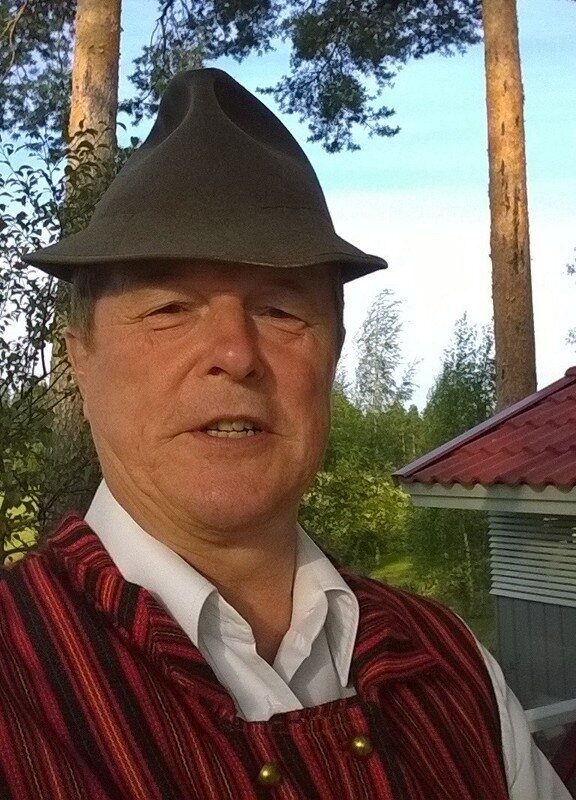 Iäkäs mies ulkona mäntymetsässä. Hänellä on päässään tumma huopahattu ja yllään valkoinen paita sekä punasävyinen raidallinen liivi. Taustalla näkyy puita ja pieni rakennus, jossa on punainen katto.