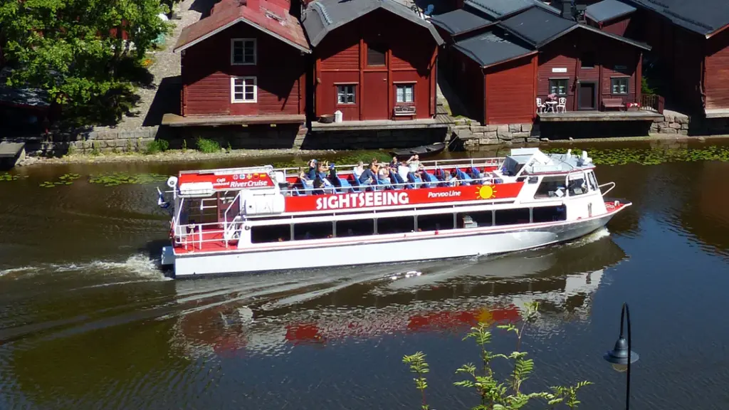 Jokiristeilyalus, jossa on matkustajia yläkannella, kulkee rauhallisesti joella. Taustalla näkyy punaisia puutaloja veden äärellä kesäisessä maisemassa.