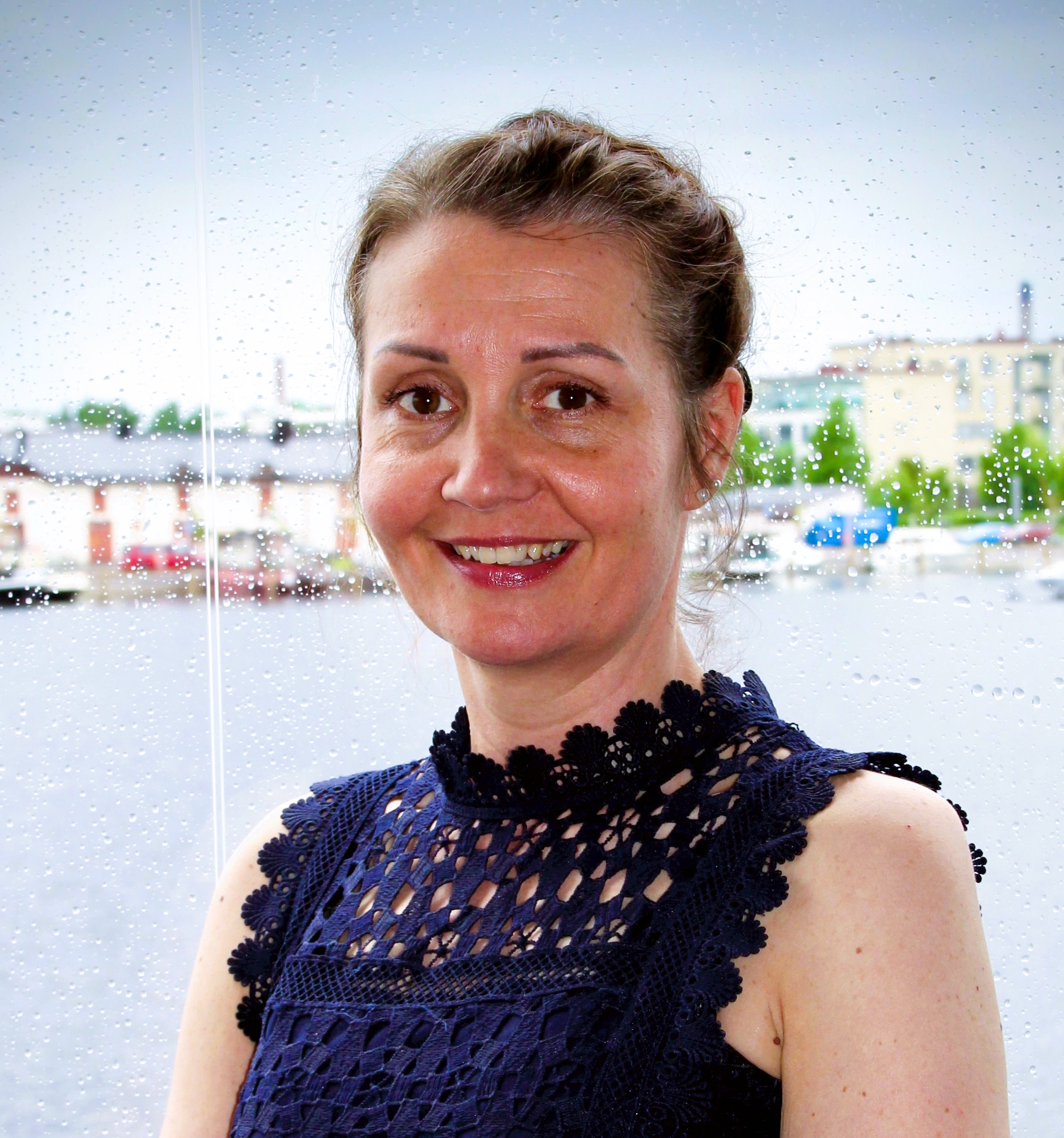 Nainen hymyilee kameralle suuren ikkunan edessä. Ikkunalasissa näkyy sadepisaroita, ja taustalla erottuu kaupunkimaisema. Hänellä on yllään tummansininen, pitsikoristeinen hihaton pusero.