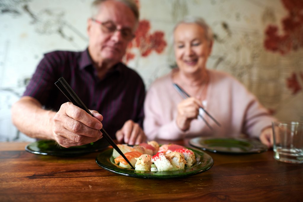 Iäkäs pariskunta istuu pöydän ääressä syömässä sushia. Mies ottaa sushipalan syömäpuikoilla ja nainen hymyilee vieressä. Pöydällä on lautasia ja juomalasi.