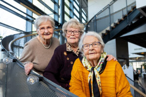 Kolme iäkästä naista seisoo sisätilassa portaikon vieressä ja katsoo kameraan. He hymyilevät ja nojaavat kaiteeseen, taustalla näkyy moderni rakennus ja kierreportaat.
