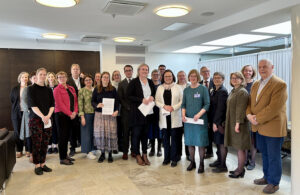 Ryhmä eri-ikäisiä asiantuntijoita ja toimijoita seisoo sisätiloissa vierekkäin, osa pitää käsissään asiakirjoja. Tunnelma on virallinen ja yhteinen, ikään kuin tilaisuuden tai tapaamisen päätteeksi otettu ryhmäkuva.