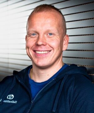 Mies hymyilee kameralle sisätilassa. Hänellä on lyhyet hiukset ja päällään tumma vetoketjullinen takki. Taustalla näkyy vaalea, vaakasuorilla säleillä varustettu seinä.