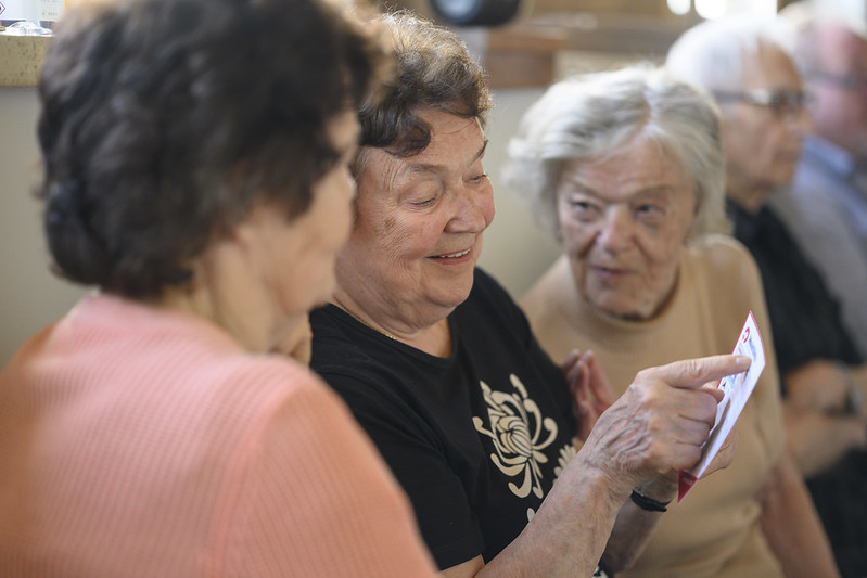Useita iäkkäitä ihmisiä istuu vierekkäin sisätilassa. Yksi naisista näyttää paperia ja osoittaa sitä muille, jotka seuraavat kiinnostuneina. Tunnelma on keskusteleva ja yhteinen.