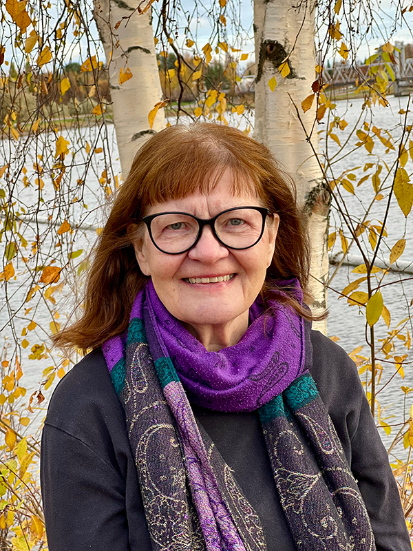 Iäkäs nainen hymyilee kameralle ulkona koivujen edessä. Hänellä on silmälasit, tumma paita ja violetti huivi. Taustalla näkyy syksyinen maisema, keltaisia lehtiä ja vettä.