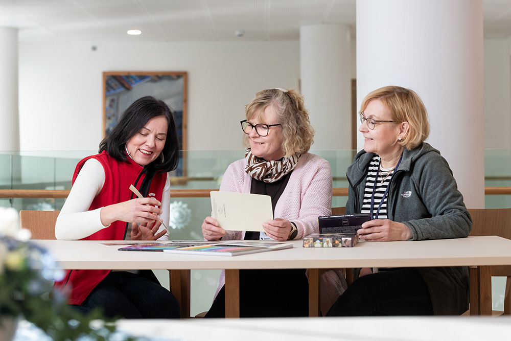 Kolme naista istuu pöydän ääressä valoisassa sisätilassa. He keskustelevat ja tekevät yhdessä jotakin luovaa – yksi näyttää muille välinettä, toinen pitää kädessään paperia ja kolmas katsoo vierestä. Pöydällä on värikyniä ja tarvikkeita.