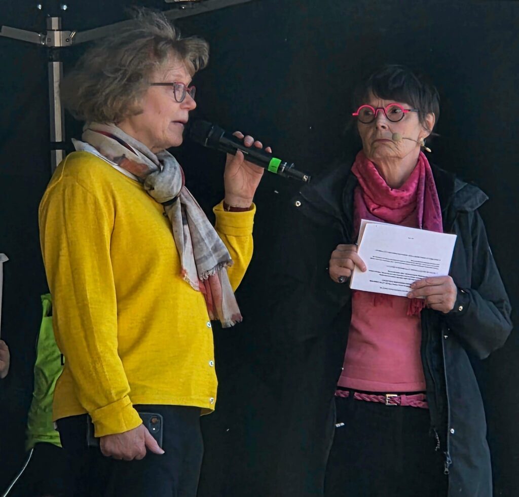 Kaksi naista seisoo lavalla mikrofonien kanssa. Toinen puhuu mikrofoniin ja toinen pitää kädessään paperia. Molemmilla on värikkäät silmälasit ja huivit, ja taustalla näkyy tumma esiintymislava.