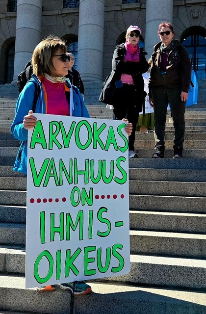 Nainen seisoo portailla kädessään suuri kyltti, jossa lukee ”Arvokas vanhuus on ihmisoikeus”. Taustalla näkyy muita tapahtumaan osallistuvia ihmisiä aurinkoisessa ulkoilmassa kivirakennuksen edustalla.
