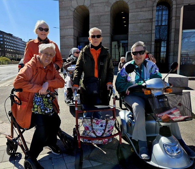 Neljä hymyilevää iäkästä naista poseeraa yhdessä ulkona Helsingin keskustassa. Kolmella on rollaattori ja yksi istuu sähkömopolla. Taustalla näkyy kivirakennus ja aurinkoinen kevätpäivä.