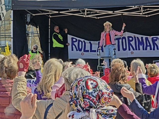 Ulkolavalla esiintyvä nainen puhuu yleisölle Rollaattorimarssin tapahtumassa. Yleisö nostaa käsiään ilmaan, ja lavan taustalla näkyy banderolli, jossa lukee ”Rollaattorimarssi”.