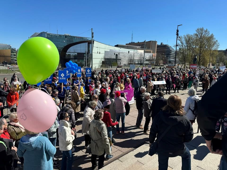 Suuri joukko ihmisiä kokoontuneena Helsingin keskustaan aurinkoisena kevätpäivänä Rollaattorimarssiin. Etualalla näkyy vihreä ja vaaleanpunainen ilmapallo sekä osallistujia banderolleineen ja kylttien kanssa. Taustalla näkyy modernia kaupunkimaisemaa ja tapahtumaan osallistuvia ihmisiä eri puolilla aukiota.